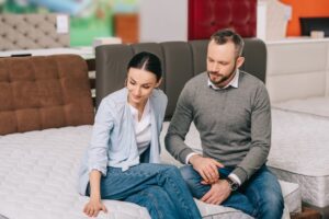 Portrait Of Couple Sitting On Mattress While Choosing Goods Together In Furniture Store