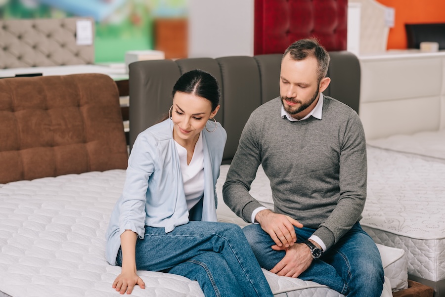 Portrait Of Couple Sitting On Mattress While Choosing Goods Together In Furniture Store